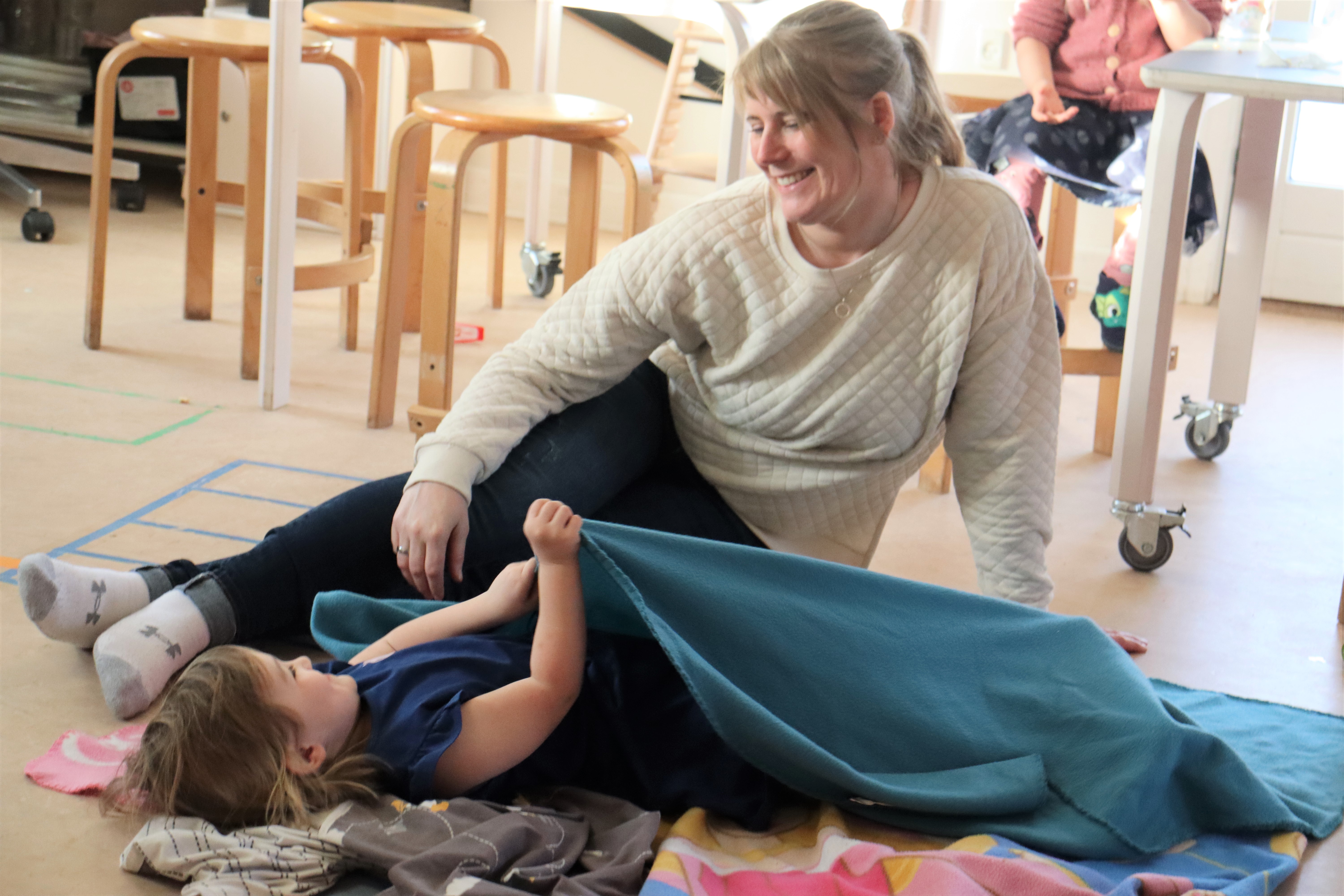 Pædagog putter barn under tæppe, hvor barnet smiler op til pædagogen 